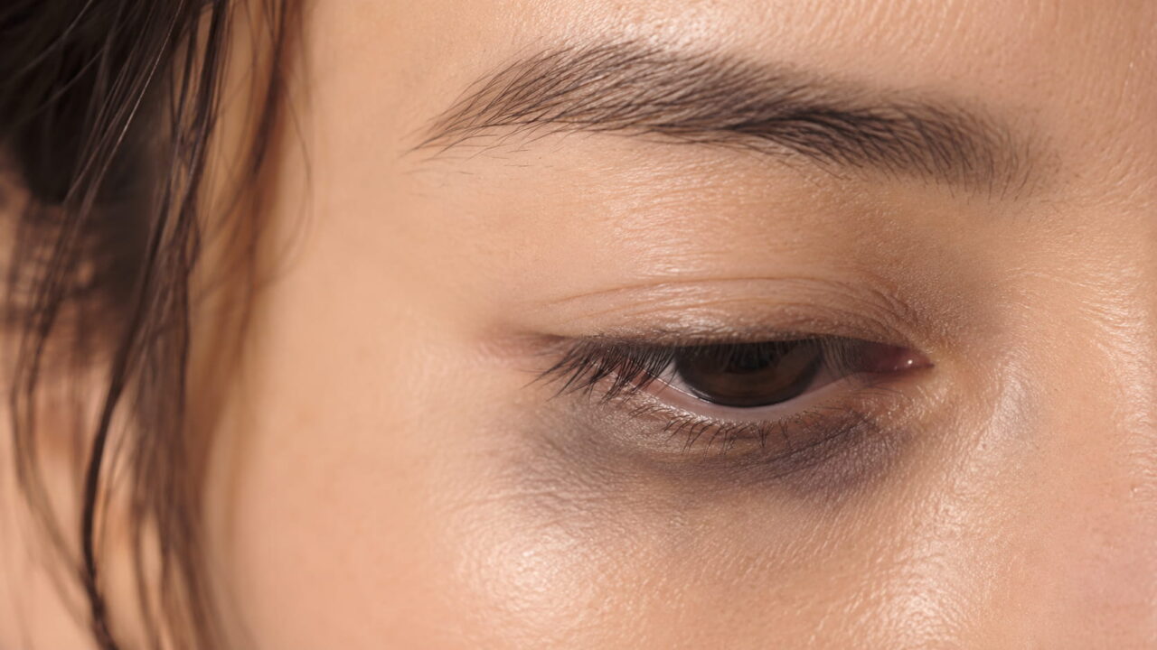 夕方の30代女性の目もとのクローズアップ。伏し目で、下まぶたに黒っぽいラインが移ってパンダ目になっている。手前に繰り出し式のペンシルアイライナー