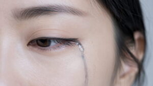 30代女性の目もとのクローズアップ。目尻から頬へ涙が伝い、その跡に沿ってアイラインが灰色ににじんで流れている