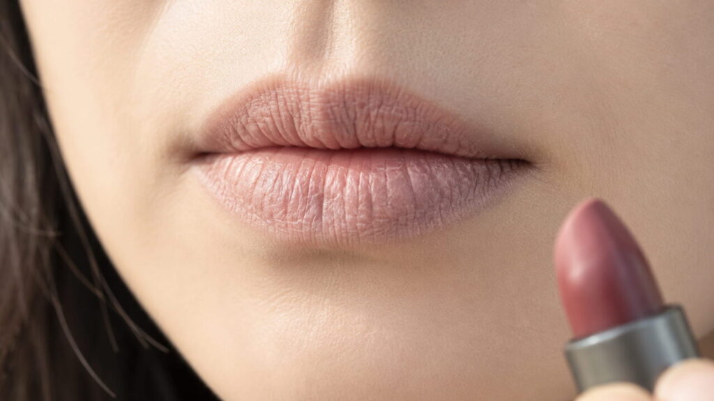 30代女性の唇のクローズアップ。口紅を塗っていない唇が乾いて縦の溝が深く刻まれ、輪郭がぼやけて口角の外の皮膚まで乾きが続いている。手前に無地のリップスティックを持つ手。リンゴ酸ジイソステアリル不使用のリップを成分表示から探す記事のイメージ